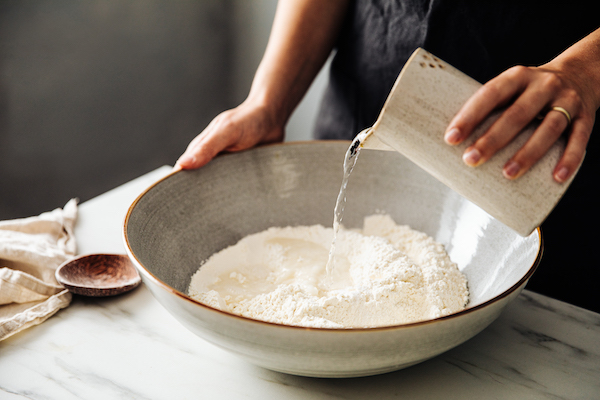 Woman pouring water on rye and wheat flour in bowl